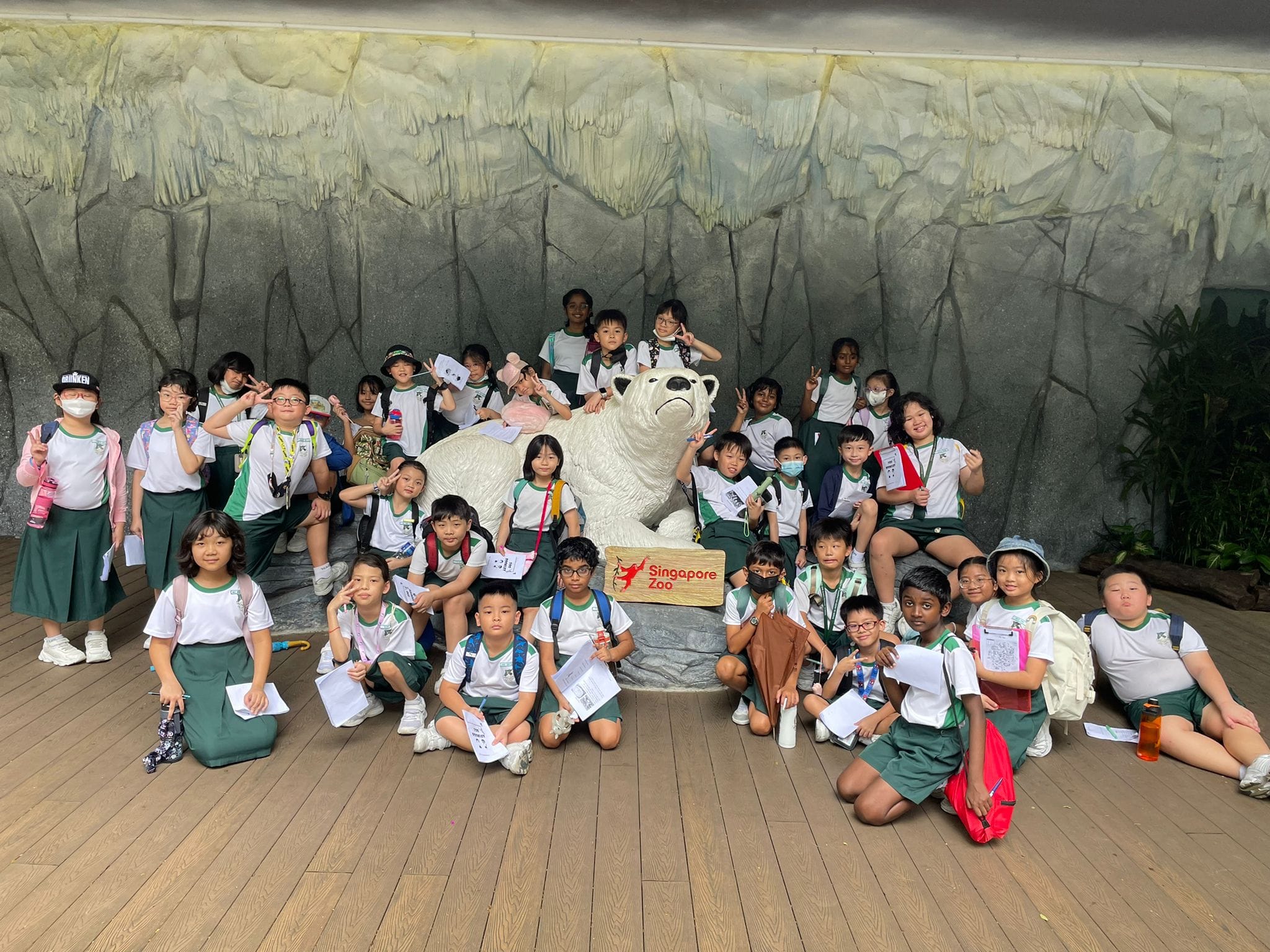 A group of young students in green and white uniforms pose around a polar bear statue at Singapore Zoo.